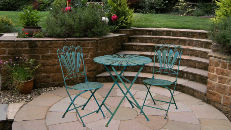 Je voulais que mon minuscule espace extérieur se sente comme un balcon parisien chic – et cet ensemble de bistrot de style vintage à 180 $ est la réponse facile Teal wrought iron outdoor bistro table and chairs sitting on a stone patio surrounded by brick planters filled with greenery