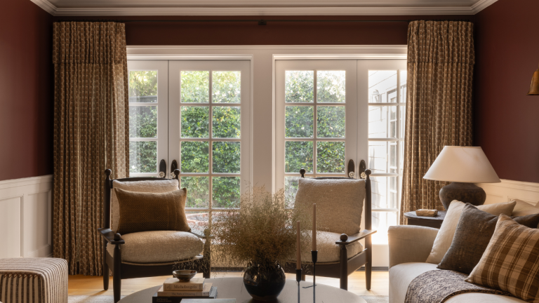 Vos fenêtres méritent également le traitement « California Cool » d’Amber Lewis – cette dernière collaboration apporte son style décédé décédé aux rideaux et aux nuances a living room with red walls, a set of two chairs, a coffee table holding books, a vase, and candle holders, and double french doors to the outdoor area framed with floor length curtains
