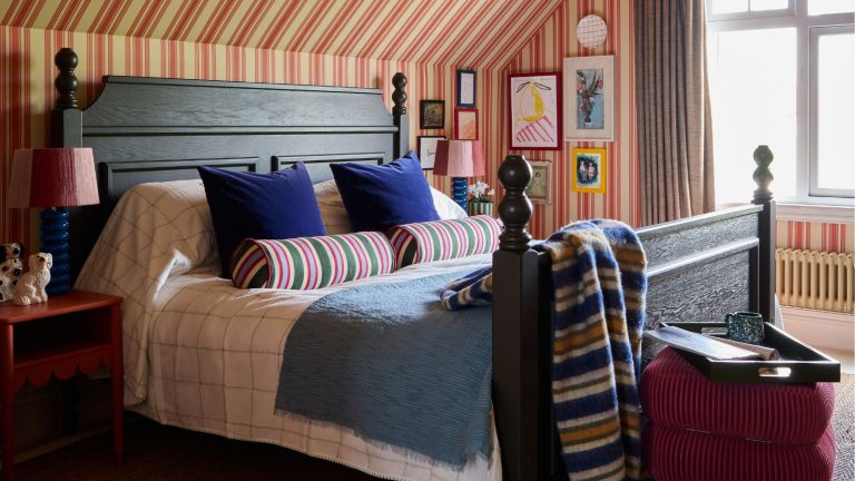 « Vous pourriez y aller un peu plus chez vous » – pourquoi ce designer pense que nous devrions tous être plus nerveux à propos de nos décisions de design d’intérieur bedroom with a sloped ceiling and beige and red striped wallpaper that goes up and over the ceiling. A large dark wooden vintage bed sits in the middle styled with striped throws and pillows with a velvet stool at the foot of the bed