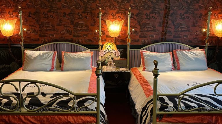 Je ne pouvais pas m’arrêter de penser aux lampes vintage de mon séjour à l’emblématique Lafayette Hotel – alors j’ai trouvé ces lookaliks tout aussi élégants pour ramener à la maison du style hollywoodien de la vieille école avec moi Eclectic guest room at the Lafayette Hotel in San Diego, featuring two four-post beds with striped headboards and pink bordered bedding, along with pink toile wallpaper and a fringe antique-style table lamp.