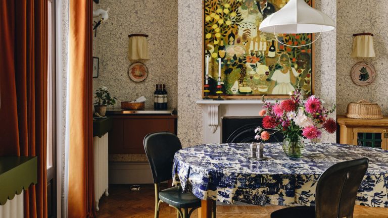 Je n’ai jamais pu décrire mon style de design d’intérieur jusqu’à ce que je tombe sur le terme maximalisme Scandi – voici tout ce que vous devez savoir sur cette tendance folky et ludique scandi maximalist style dining room with wallpapered walls, a large fireplace with folk art hung above, dark orange velvet curtains, a dining table with blue and white printed tablecloth and a white fabric pendant light