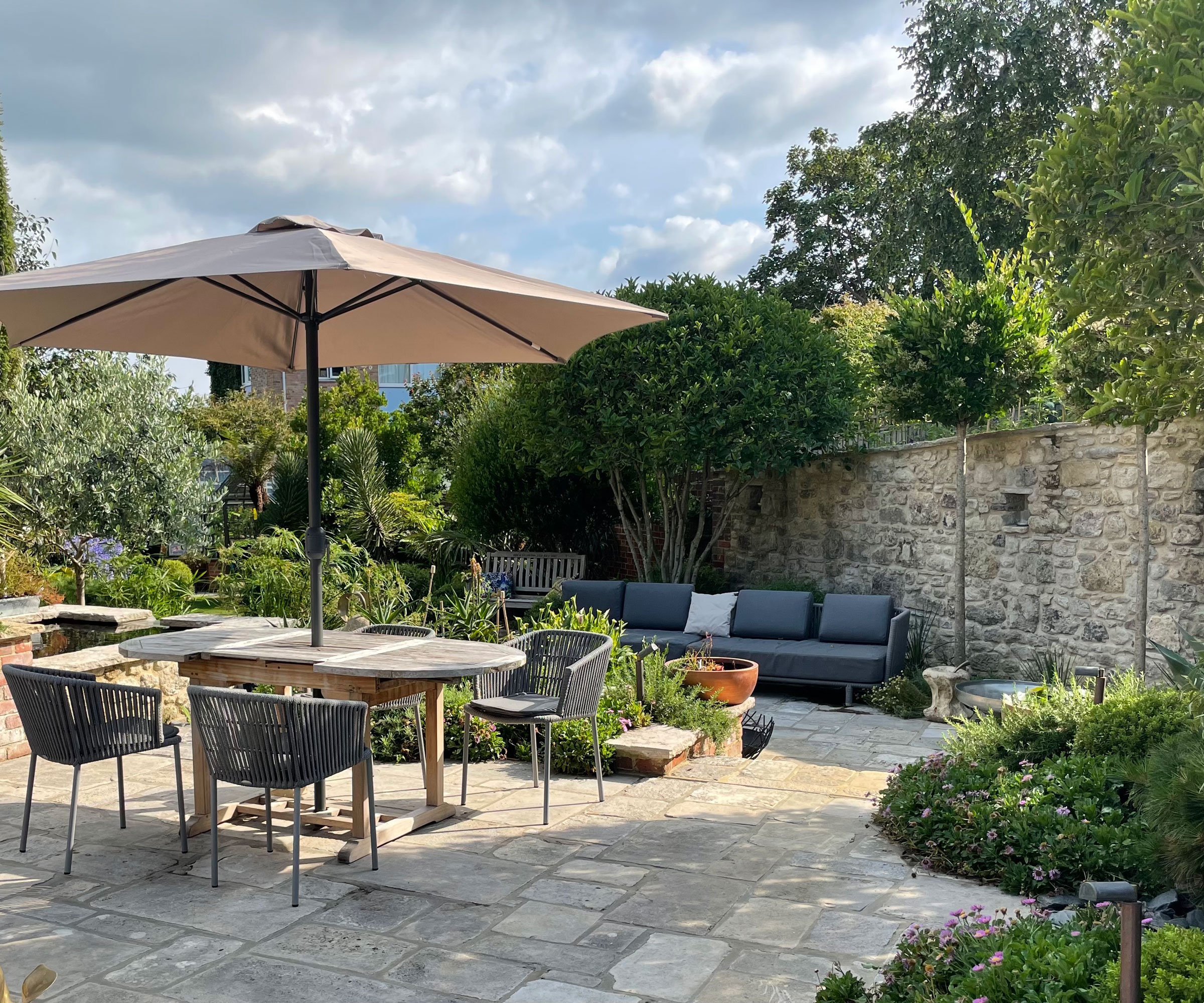 Jardin clos avec mur de briques, carrelage et mobilier de jardin comprenant un ensemble de repas, un parasol et des canapés, avec des plates-bandes et un arbre à feuilles persistantes à plusieurs tiges