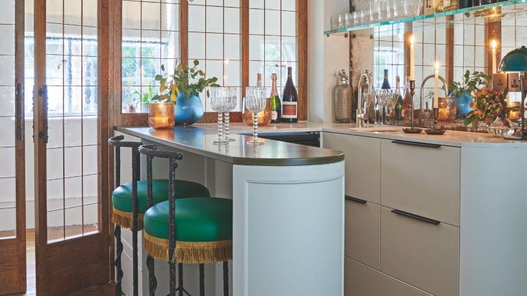 Cette minuscule étagère chromée de 20 $ a donné à mon appartement le micro-bar de style parisien qu’il a manqué – tous mes invités adorent cette nouvelle fonctionnalité Corner home bar with a peninsular layout, pale blue cabinets, dark stone top, green vintage bar stool and open shelving for drinks and glasses