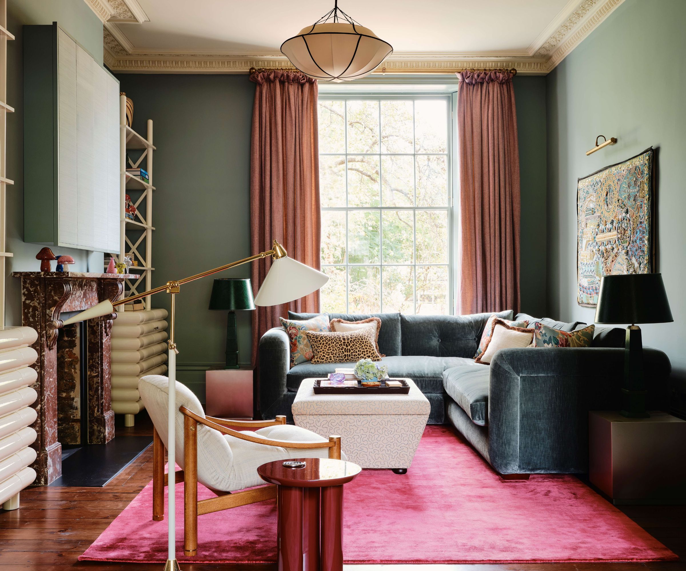salon bleu avec des rideaux roses et un tapis rose avec un canapé d'angle en velours bleu profond, un pouf, un fauteuil en bois et une table d'appoint avec lampe