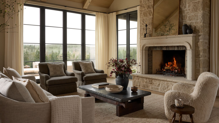 A carpeted living room with large floor-to-ceiling windows and a fire roaring in a stone fireplace with an ornate mirror resting on the mantel above. Cozily upholstered accent chairs, a sofa, and low slung charcoal coffee table holding collected objects sit before it.