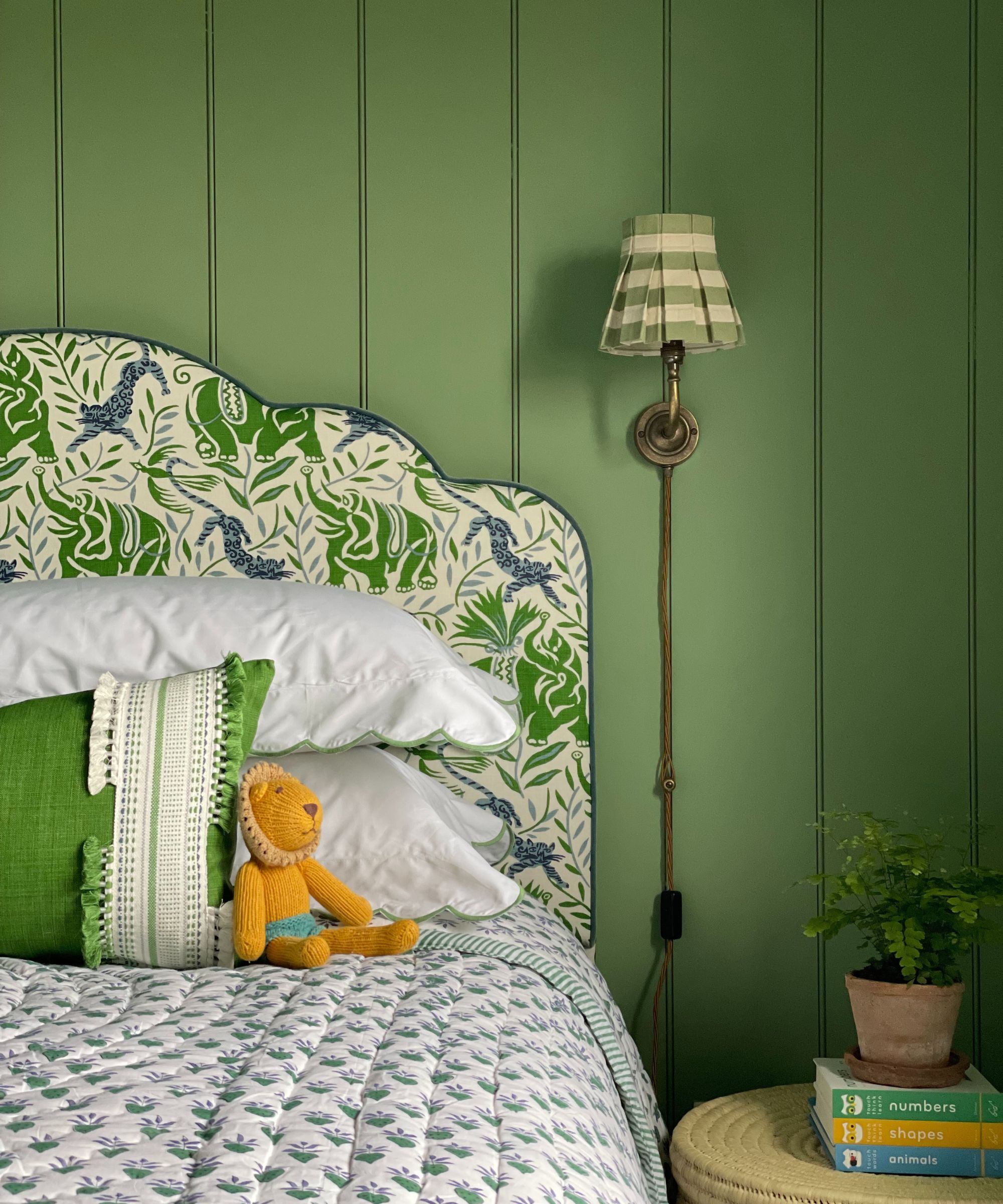 Chambre avec murs verts en lambris peints, tête de lit rembourrée avec lancer floral, table d'appoint en rotin et Scocne plissée
