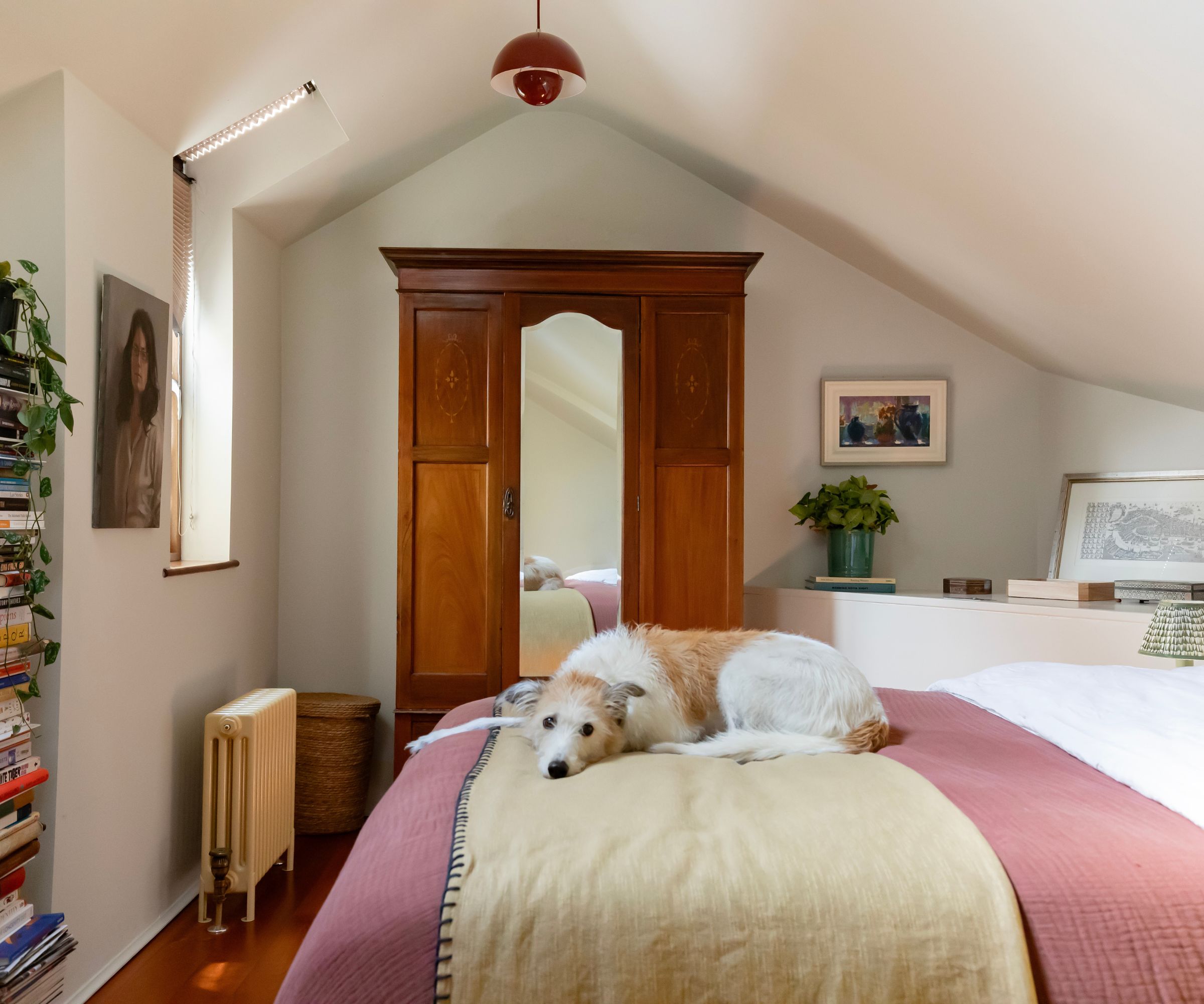 Chambre avec plafond en pente, murs blancs, garde-robe en bois foncé, lit avec lancer rose et chien
