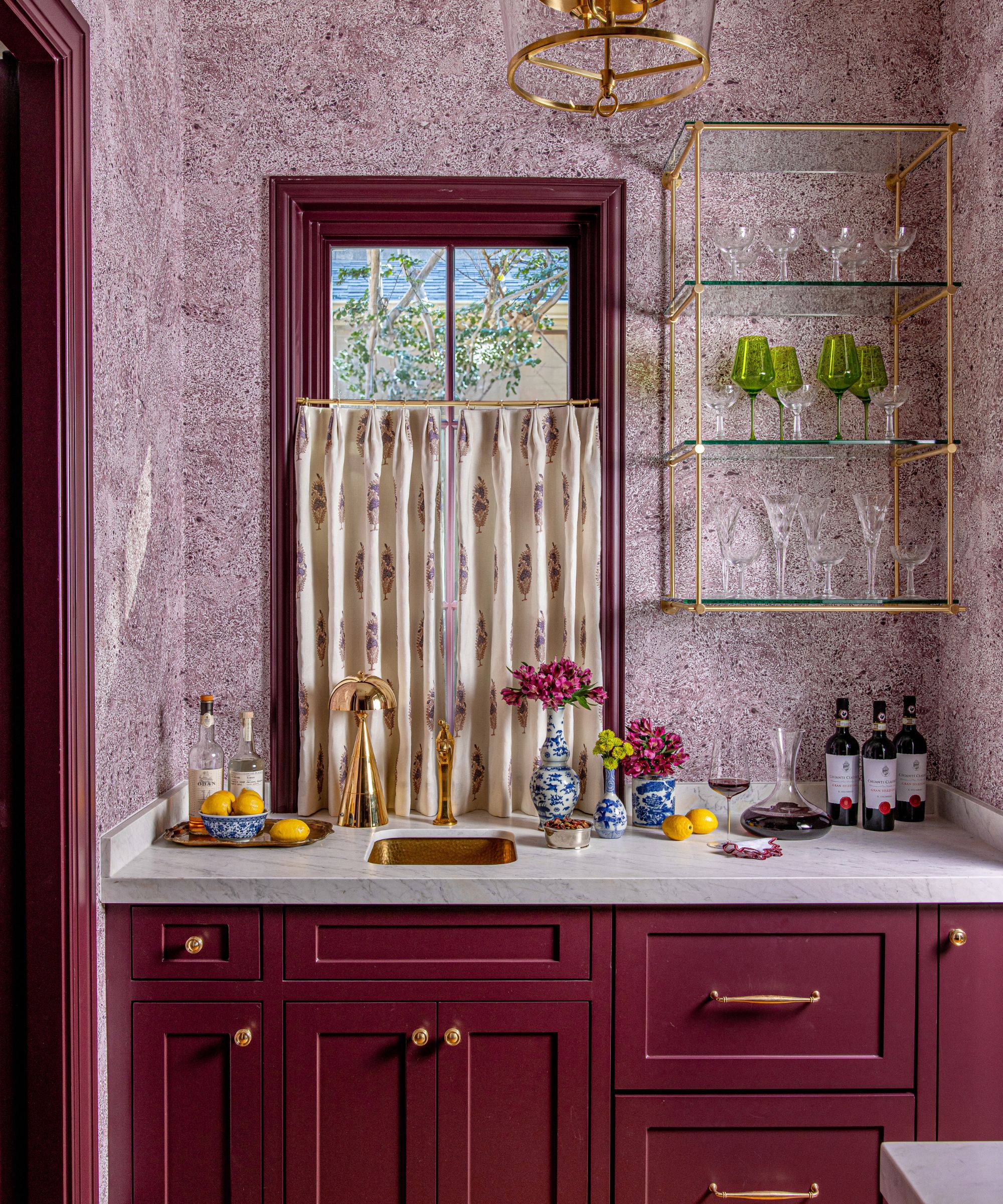 Bar à domicile avec armoires peintes en bordeaux et cadre de fenêtre, quincaillerie en or, comptoir en marbre et évier et étagères ouvertes avec des lunettes