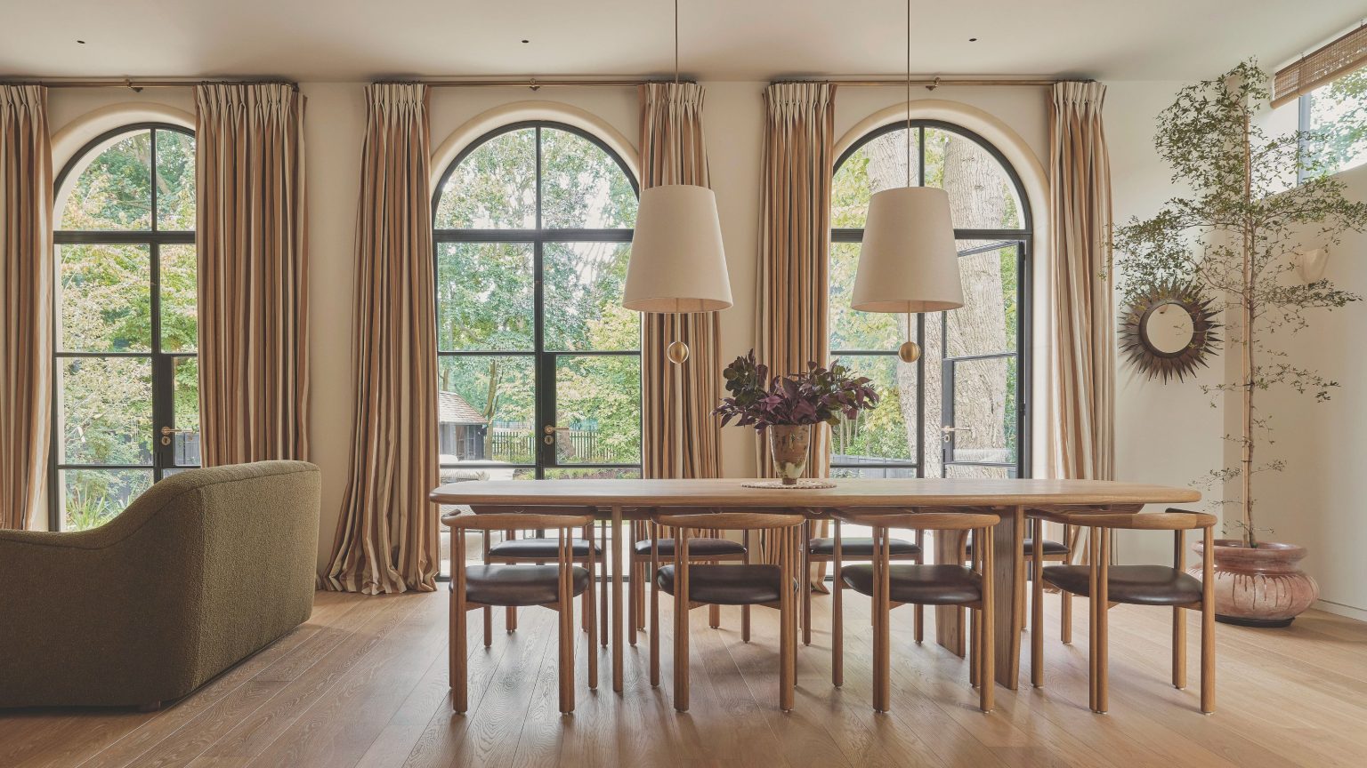 « Il aura toujours une atmosphère chaude et automnale » – explorez une élégante maison en briques rouges qui est une leçon pour créer des intérieurs confortables mais chics Open plan dining room with three large arched windows each with long cream drapes. A large wooden dining table sits in the middle of the space with two pendant lights with white shades hanging above