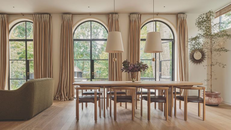 « Il aura toujours une atmosphère chaude et automnale » – explorez une élégante maison en briques rouges qui est une leçon pour créer des intérieurs confortables mais chics Open plan dining room with three large arched windows each with long cream drapes. A large wooden dining table sits in the middle of the space with two pendant lights with white shades hanging above
