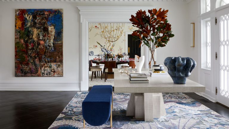 Comment concevez-vous une maison vibrante lorsque le propriétaire est daltonien? Pour ce designer, la création de pièces qu’ils pouvaient «ressentir» sont devenues le défi le plus inspirant Large entryway with white walls and a wooden floor. A stone table sits in the middle decorated with books and a large vase of foliage. There is a blue bench next to the table and the both sit on a patterned blue and white rug