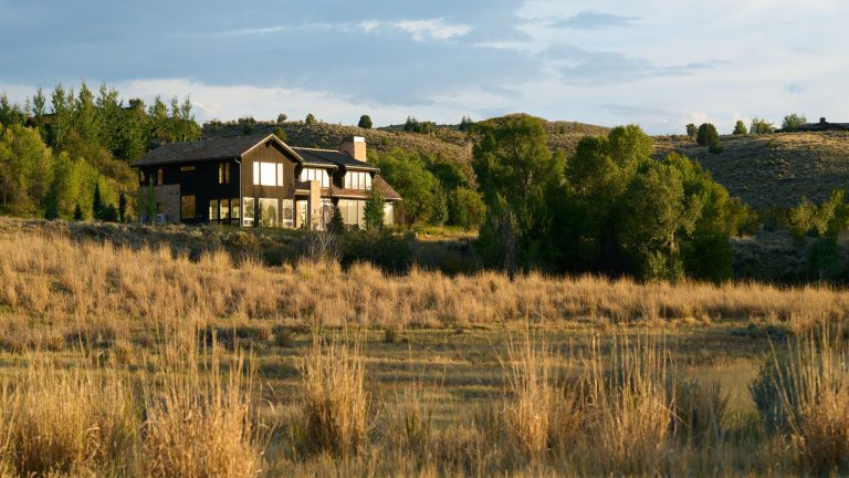 S’assurer que cette maison de nouvelle construction était silencieusement assis parmi son environnement étonnant était le facteur le plus important de ce projet collaboratif A modern stone and wood house surrounded by tall grasses and trees, set against rolling hills at sunset
