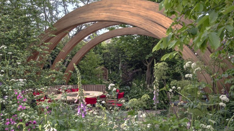 5 tendances inspirantes en matière d’aménagement paysager qui définiront 2026 – et pourquoi les concepteurs disent qu’ils sont là pour rester A show garden at Chelsea Flower Show with trees, shrubs, and flowering perennials under a large wooden arch