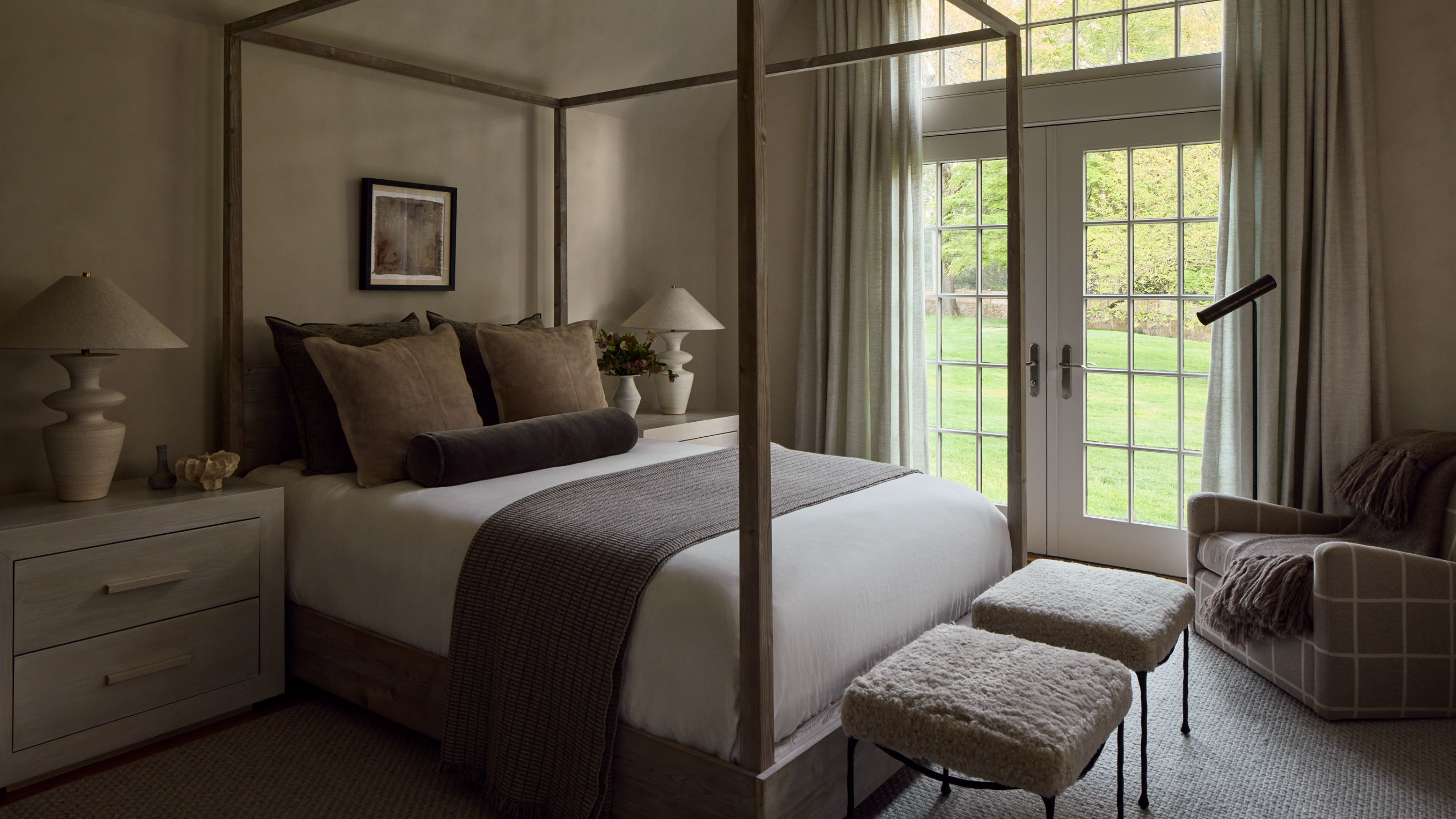 Chambre neutre avec lit à baldaquin et grandes portes-fenêtres. Il y a de longs rideaux autour des fenêtres et le lit est décoré de plusieurs draps neutres.