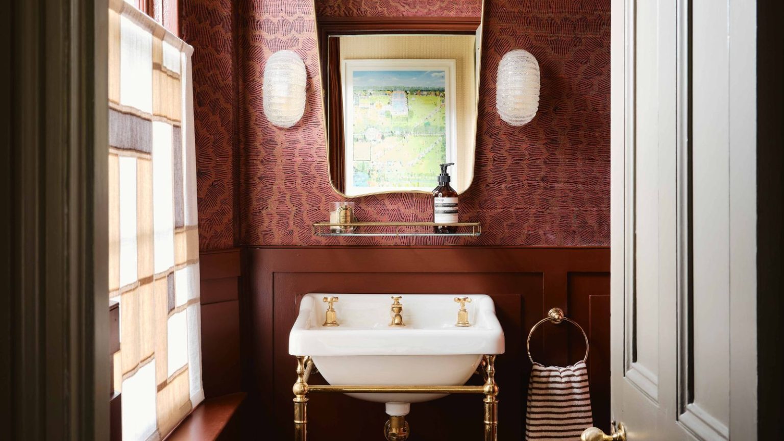 dark burgundy red powder bathroom with panelled walls and wallpaper above, with a mid century mirror, vintage wall sconces, and a traditional sink on a brass stand