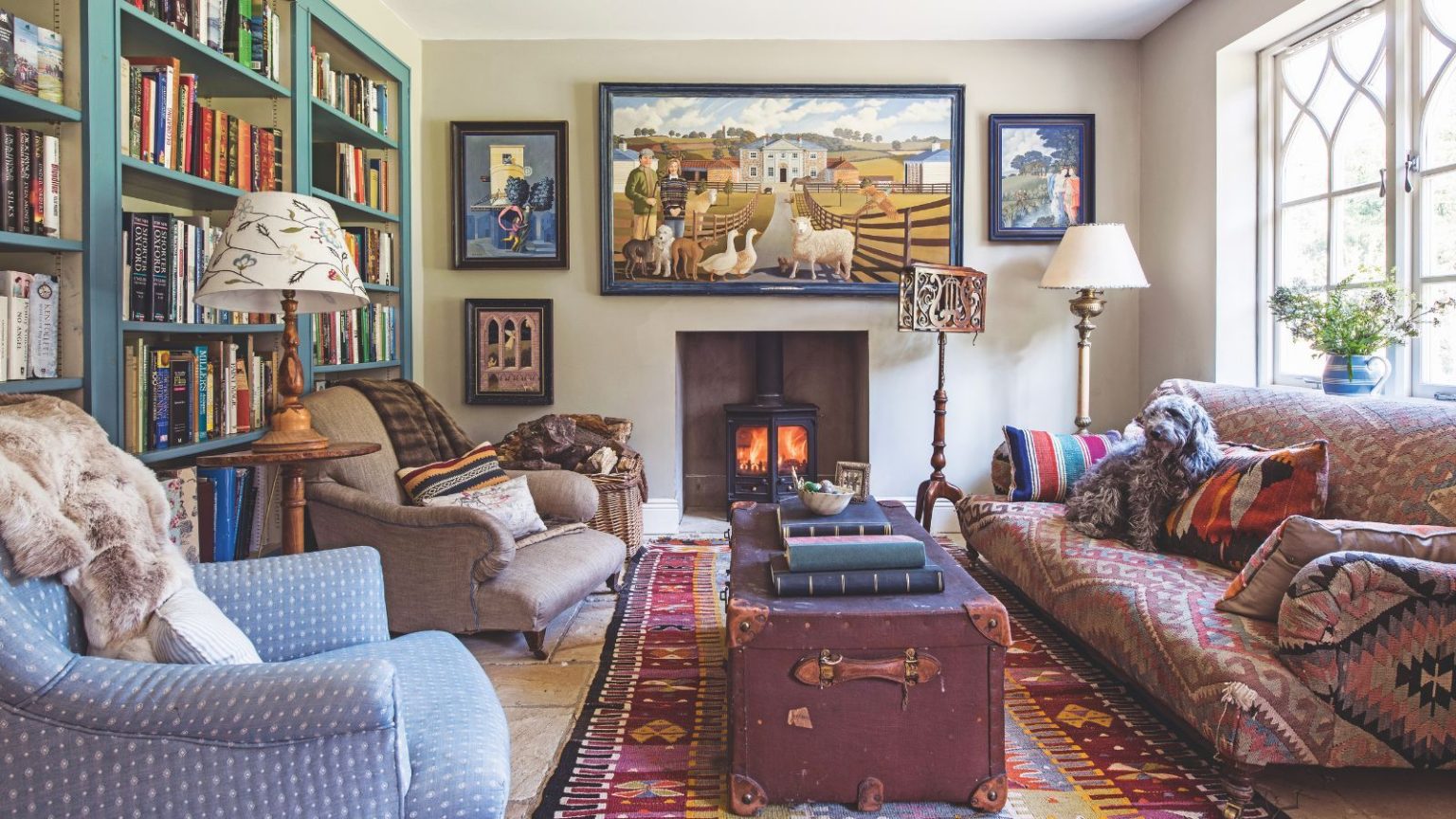 3 essentiels du Feng Shui d’automne pour rendre votre maison plus confortable instantanément – les thérapeutes d’intérieur jurent qu’ils vous aideront à vous détendre facilement A cozy and eclectic living room with a library wall, lit wood burner, patterned large rug, vintage look trunk and a mismatched upholstered sofa and chairs.