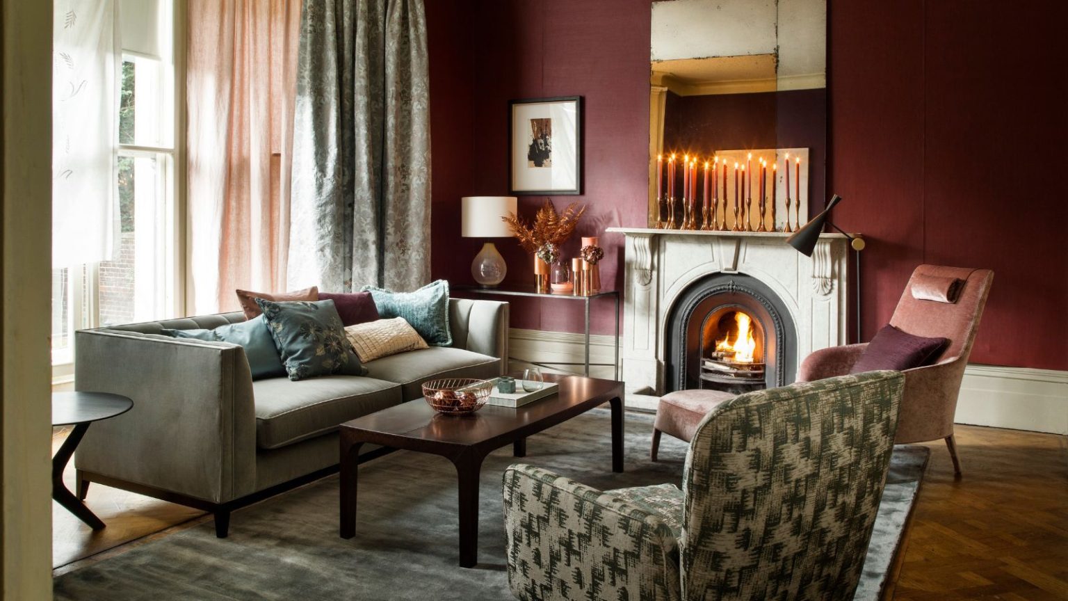 « Incroyablement facile à installer » : cet ensemble de qualité de l’air pour maison intelligente est essentiel pour éviter les maladies pendant la période des fêtes – des milliers de personnes l’ont acheté le mois dernier seulement A cozy living room with burgundy walls, mismatched upholstered furniture, a large grey velvet rug, brown wooden coffee table, and fire burning in a stone fireplace with tall tapered candles burning on the mantelpiece above.