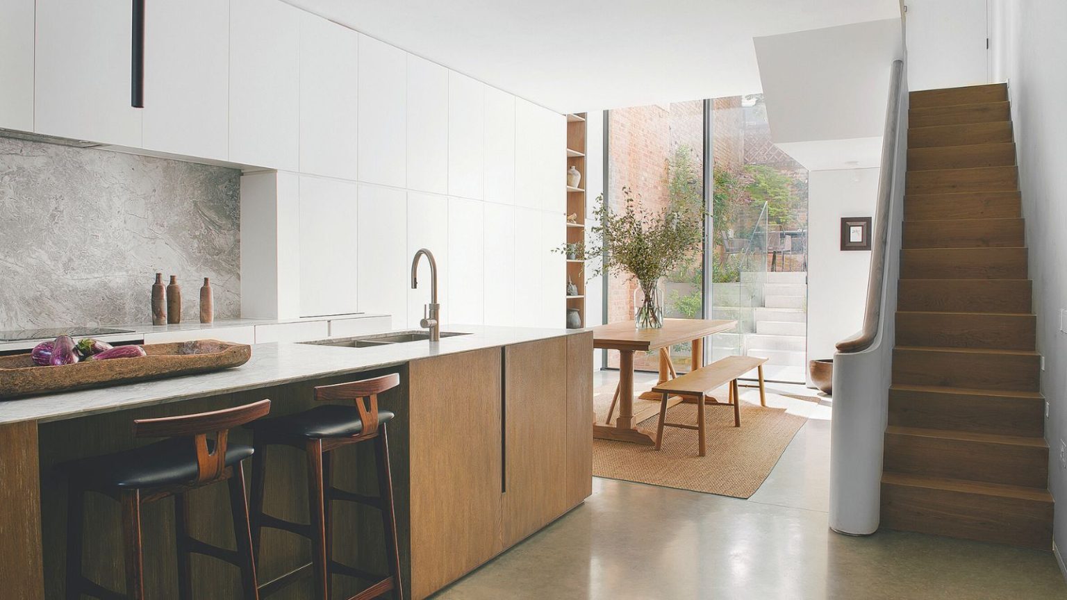 La solution non toxique, durable, bon marché et simple que les propriétaires peuvent utiliser pour éloigner les souris en hiver et au-delà A white kitchen with a wooden kitchen island with in-built sink. A staircase on the right hand side leading up, and a wooden dining table and benches beside it. Large floor-to-ceiling glass at the back of the room showing a small patio.