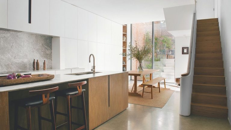 La solution non toxique, durable, bon marché et simple que les propriétaires peuvent utiliser pour éloigner les souris en hiver et au-delà A white kitchen with a wooden kitchen island with in-built sink. A staircase on the right hand side leading up, and a wooden dining table and benches beside it. Large floor-to-ceiling glass at the back of the room showing a small patio.