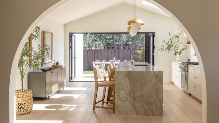 Arrêtez de respirer la poussière – Ce puissant aspirateur Miele avec sac a réduit mes allergies (et dure 20 ans) Large kitchen and dining area with curved archway, marble kitchen island, countertop stools, hanging lights, large plant in wicker pot, and large, open window revealing outdoor area.