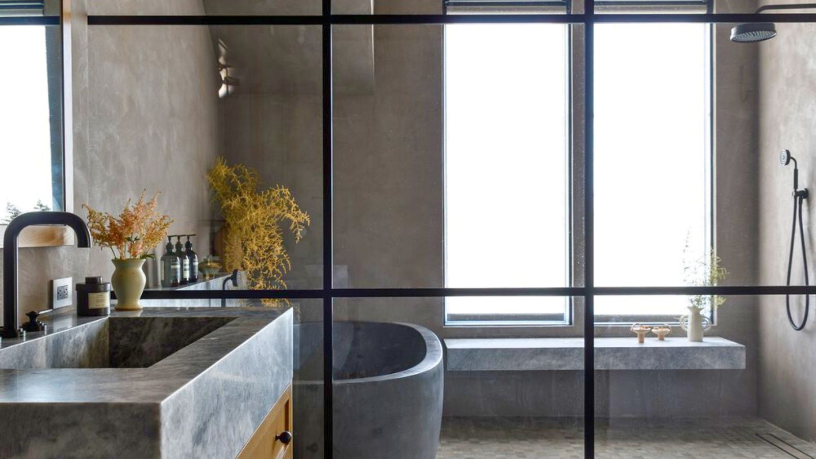 Douche en verre Crittal dans salle de bain en pierre neutre. Il y a un siège de douche et une grande fenêtre dépolie visibles, ainsi qu'une baignoire et un lavabo en pierre grise.