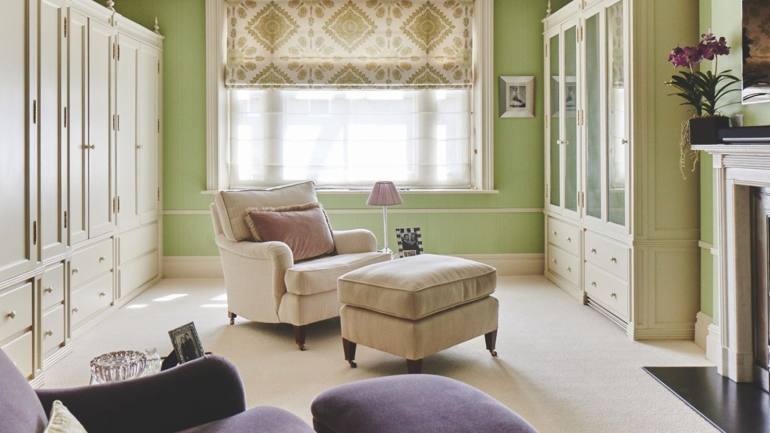 J’ai essayé la règle de désencombrement un sur deux – cela a empêché ma maison de sombrer dans le chaos cette saison des achats A green dressing room with white carpet, white built-in wardrobes, a large window with a sheer curtain and green and white patterned blind, and a white fireplace to the right. In the center of the room is a white upholstered chair with matching footrest and pink lamp and cushion, and in the foreground is a purple upholstered chair.