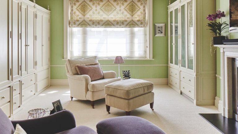 J’ai essayé la règle de désencombrement un sur deux – cela a empêché ma maison de sombrer dans le chaos cette saison des achats A green dressing room with white carpet, white built-in wardrobes, a large window with a sheer curtain and green and white patterned blind, and a white fireplace to the right. In the center of the room is a white upholstered chair with matching footrest and pink lamp and cushion, and in the foreground is a purple upholstered chair.