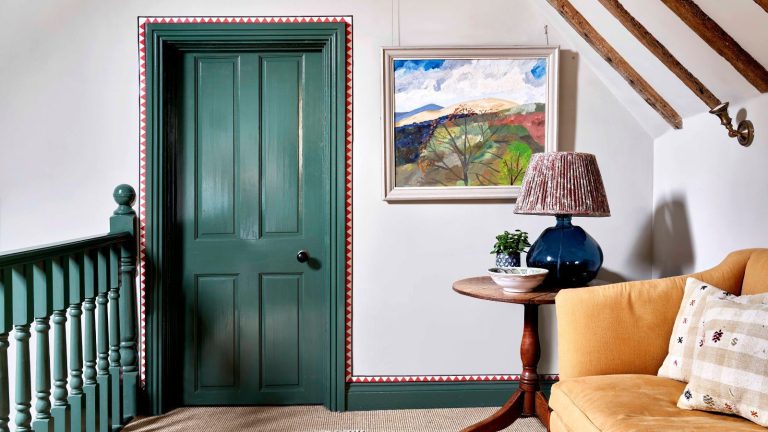A landing at the top of a staircase with green handrails, and matching green door and trim. Red detailing painted around the trim. A yellow sofa on the right hand side, next to a round wooden side table with a lamp on it.