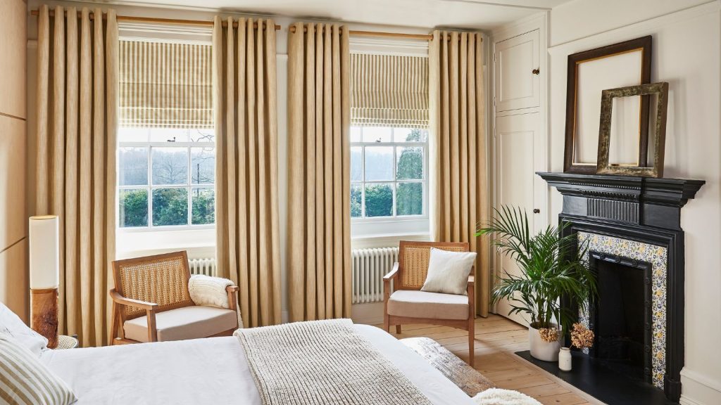 A neutral bedroom with beige curtains and blinds. Two rattan arm chairs face the bed, and a black fireplace opposite.