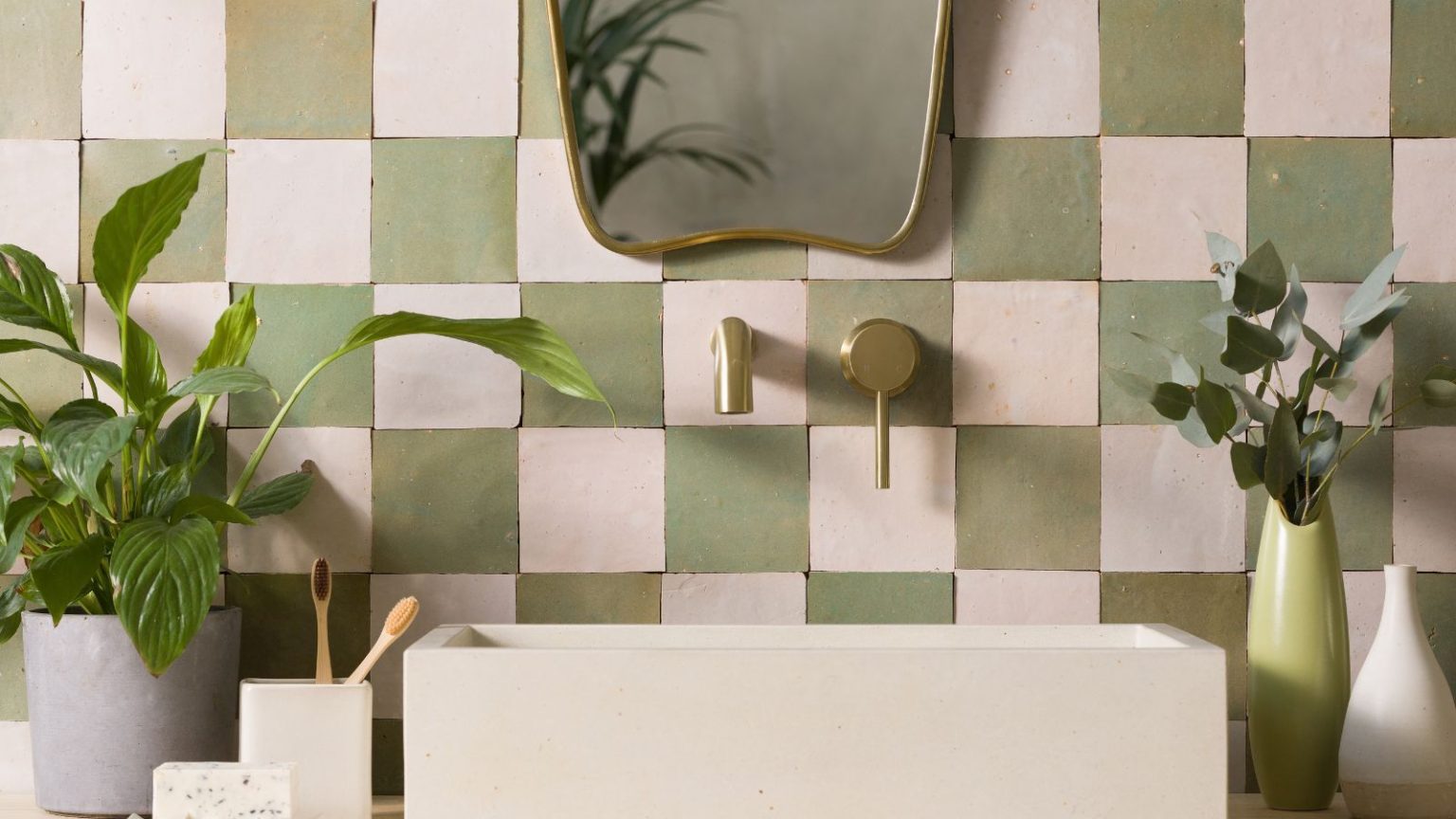 L’astuce que les experts en parfums utilisent pour donner instantanément à une salle d’eau une odeur luxueuse pour les invités d’automne – et cela dure des jours A white and green Zelige tiled backsplash wall with a white sink, gold faucets, and a gold mirror above. To either side are green houseplants.