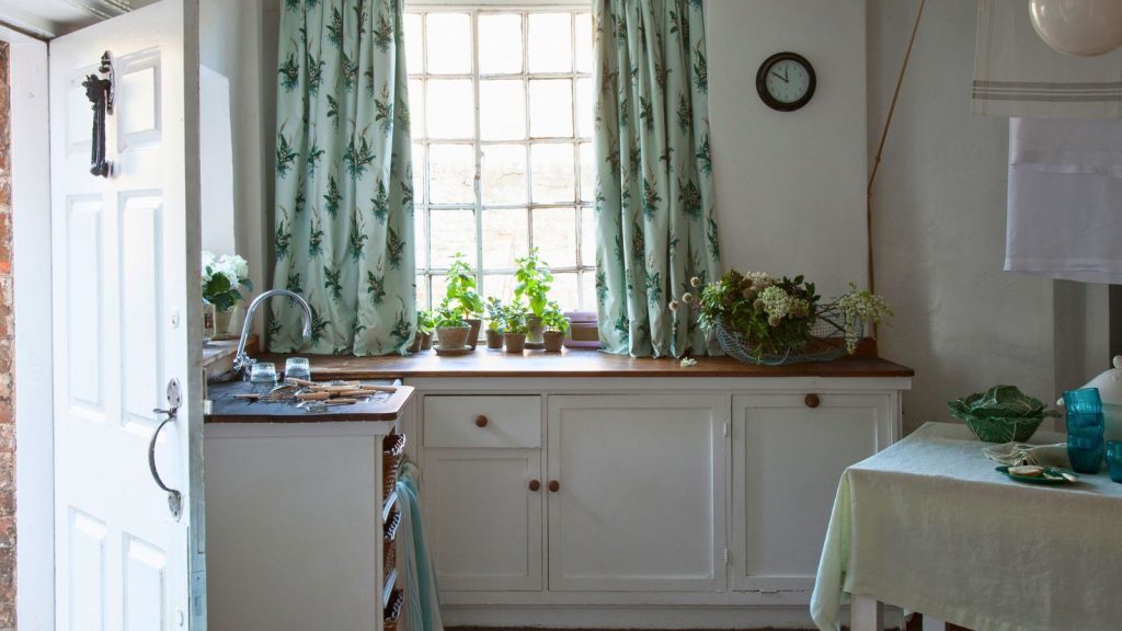 « Ramasse la poussière comme un aimant » – Scrub Daddy n’est pas seulement idéal pour les éponges, ses chiffons réutilisables, durables et lavables à 10 $ ont ravi les acheteurs A white vintage style kitchen with wooden countertops, green curtains, white walls, a black clock and dining table covered in a green tablecloth. To the left is an open white front door.