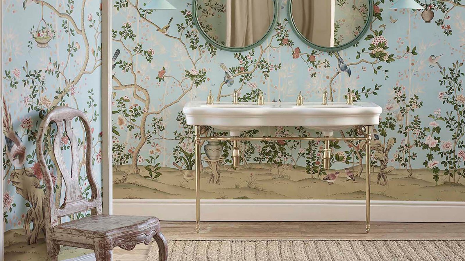 Grande salle de bains lumineuse avec papier peint bleu pâle, double lavabo victorien avec quincaillerie dorée et chaise vintage en détresse
