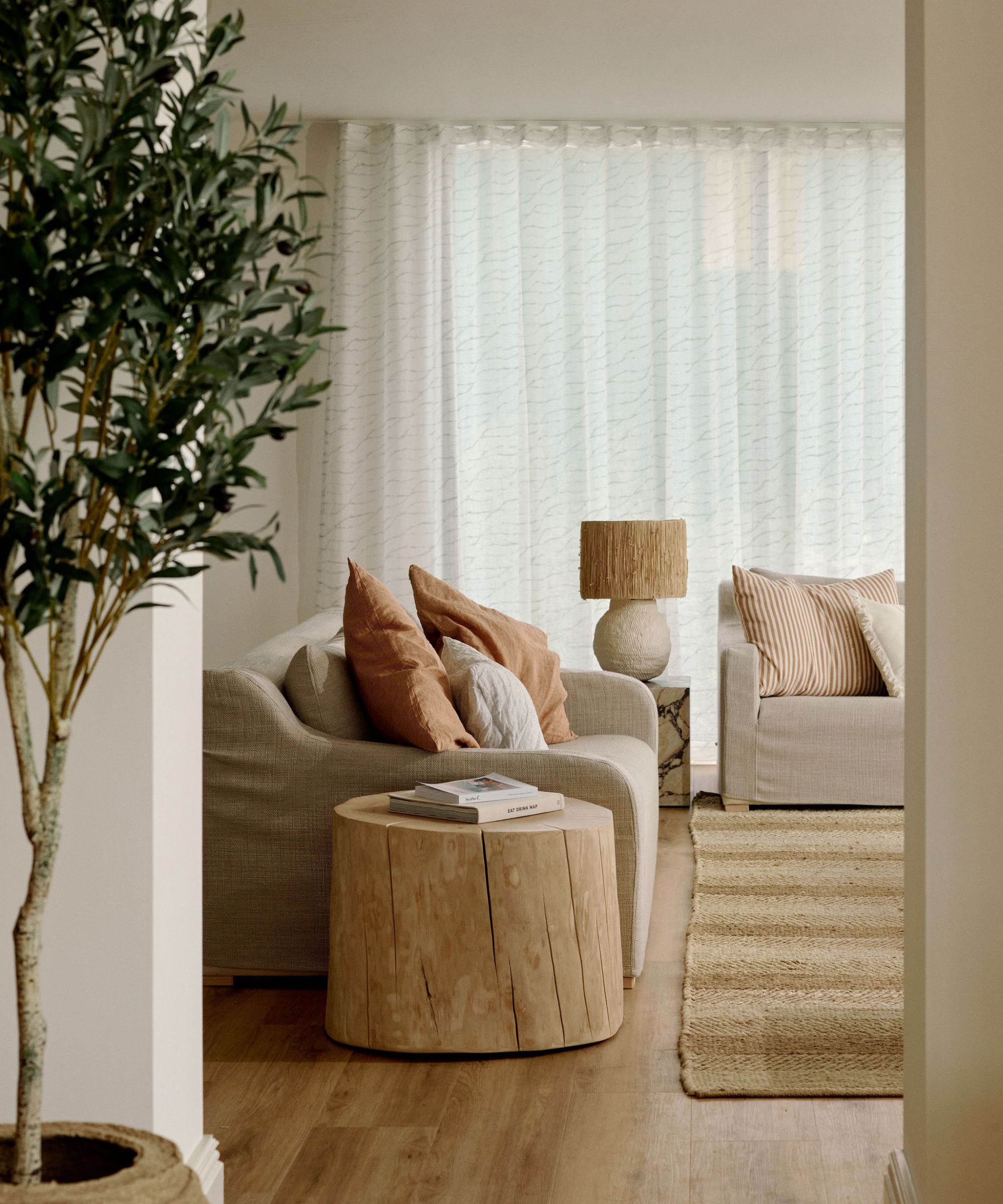 Salon minimaliste et chaleureux avec un tapis en jute, deux canapés recouverts de lin, une table d'appoint rustique en bois et un olivier