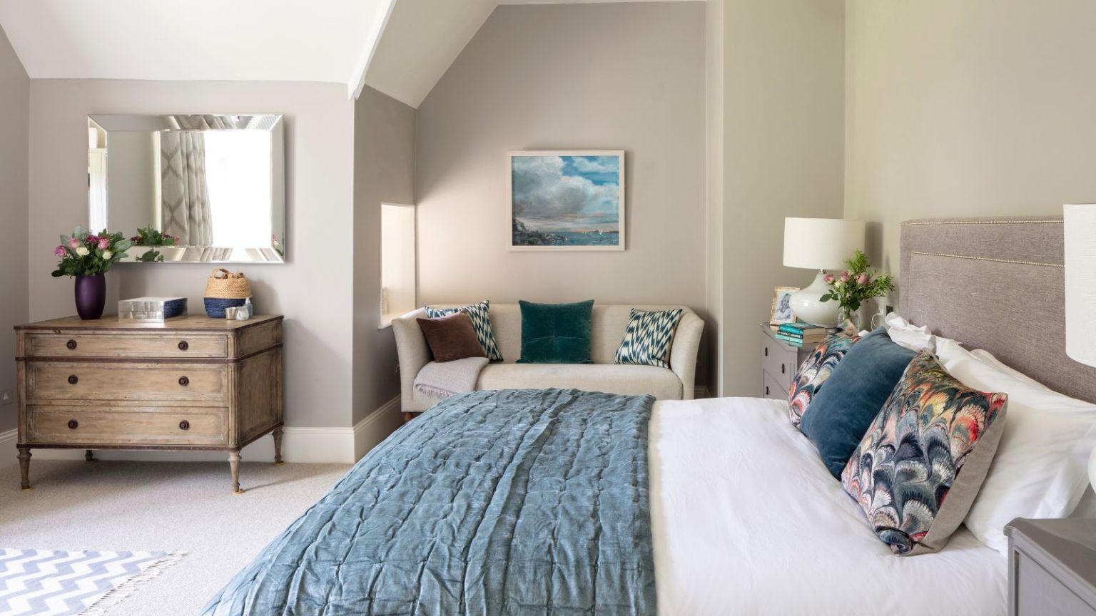A neutral bedroom with a large double bed with white and blue bedding, cushions, and matching bedside tables with white lamps. Behind is a cream colored couch with cushions beneath a framed artwork, and to the right is a brown chest of drawers with a vase of flowers on top beneath a large rectangular mirror.