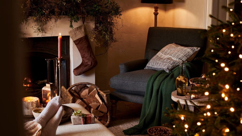 A cozy living room decorated for Christmas. Close up of a blue upholstered arm chair beside a fireplace with a stocking hanging from a garland on the mantel. The edge of a tree in the foreground.