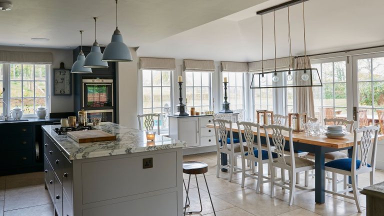 A white kitchen with marble countertops, bar stools, a large dining table with white kitchen chairs, pendant lamps, and large windows all the way round with neutral blinds.