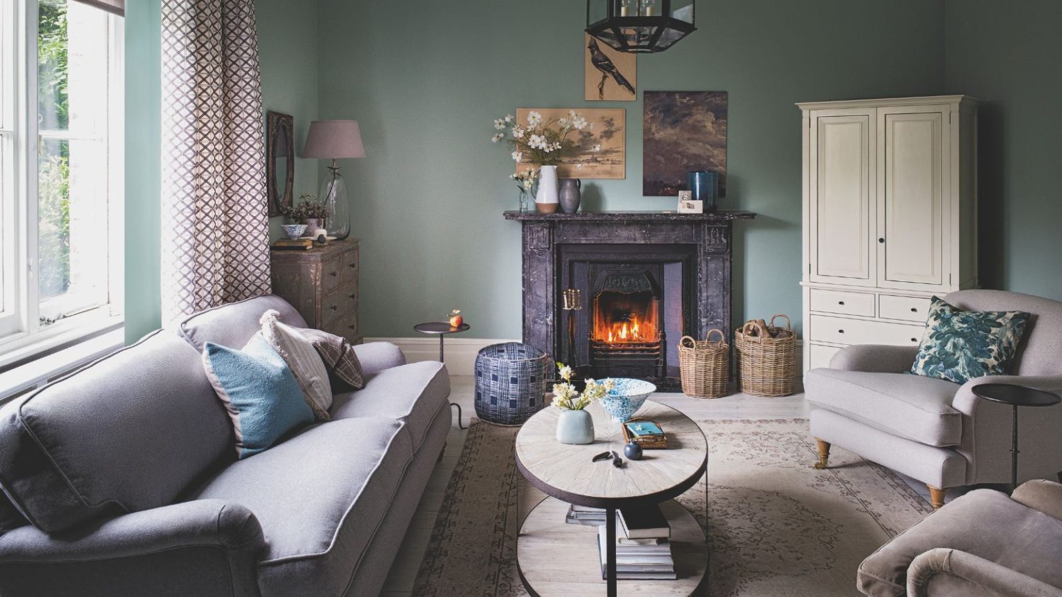 C’est le 1 article vital que les experts recommandent aux propriétaires d’avoir à portée de main alors que les avertissements de froid extrême arrivent – Restez au chaud sans faire fonctionner le chauffage en permanence. A teal living room with a grey couch, large Persian rug, coffee table, two matching grey upholstered chairs, a white closet, and a fire lit in a wood burner under a marble mantelpiece. Above are three mismatched artworks. To the left is a large window with open patterned red and white curtains.