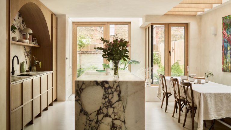 An airy kitchen diner with a large marble island separating worksurfaces and a rectangular dining table with three chairs pulled up to it.