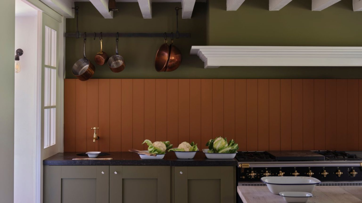 Faites briller les casseroles sales en acier inoxydable comme neuves avant les fêtes de fin d’année avec ce nettoyant « magique » le plus vendu à 7 $ que les acheteurs adorent Dark green kitchen with dark orange wall panels and copper pans hanging on rail over a small sink. The counters are black, cabinets dark green, there is a window and door to the left, and shallow dishes with fresh produce by the sink