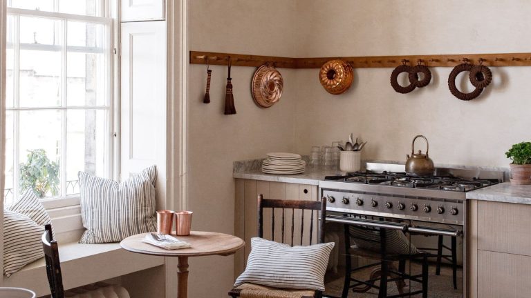 Toutes les cuisines les plus charmantes que je vois en ce moment ont en commun cette antiquité bon marché A warm neutral kitchen with wooden wall pegs displaying copper molds and a cozy eat-in area below the window with a small banquette, a round table, and two wooden chairs