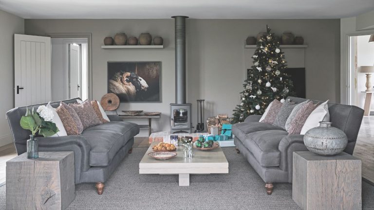 Je suis un nettoyeur professionnel et voici les 3 choses embarrassantes que je remarque toujours à propos des canapés sales lorsque je visite à Noël – et les solutions rapides A grey living room with grey couches, walls, carpet and vases. Between the couches is a white coffee table, and behind is a wood burner. To the right of the burner is a large decorated Christmas tree, and to the left is an artwork of a ram and an open doorway.