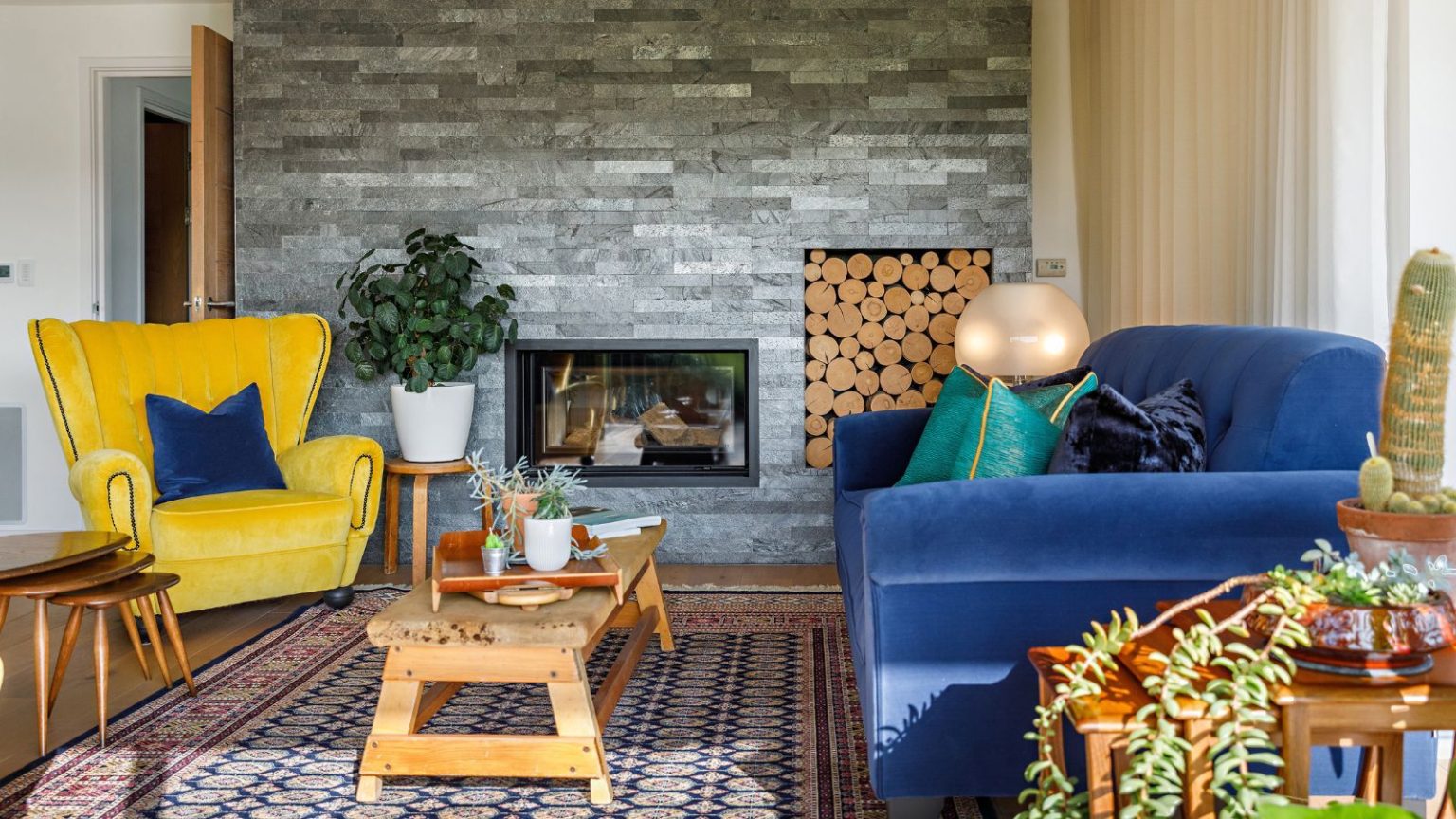 Oubliez le Black Friday, le puissant aspirateur robot de Dyson est à son prix le plus bas jamais vu – c’est l’un des plus puissants du marché pour un nettoyage vraiment sans effort Colorful, vibrant living room with blue sofa, yellow armchair, wooden coffee table, patterned rug and plants, pictured in front of gray brick-style wall, fireplace and built-in log storage.