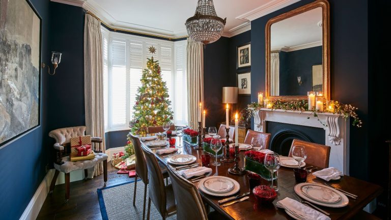 6 essentiels pour donner à votre maison une odeur de Noël – avec de la verdure luxueuse, des épices et des notes au coin du feu A blue dining room decorated for Christmas with a large laid table, large Christmas tree, presents, taper candles and a decorated white mantelpiece with candles, foliage, and a large gold mirror above.
