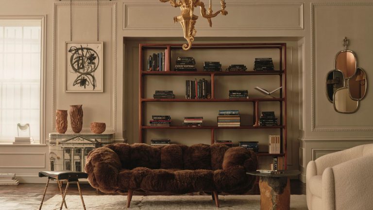 Cream living room with paneled walls and modern furniture, including a brown furry couch, a wooden minimalist bookcase and a cream boucl&amp;eacute; arm chair