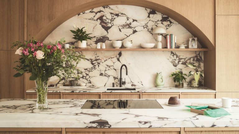 A modern kitchen with arched lightwood fittings over a kitchen sink, with a large marble backsplash. An island opposite with matching marble top, and a glass stove top. A vase of roses to the left.