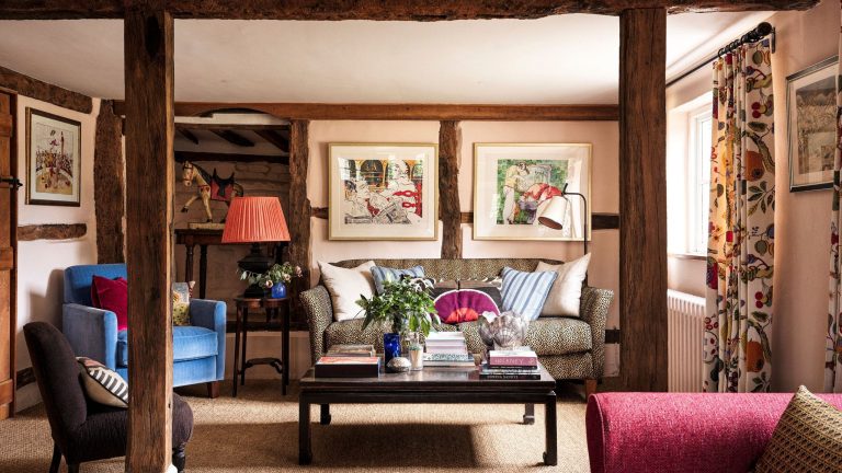 Le chalet confortable de la designer Sophie Garland ressemble à quelque chose directement sorti d’un film de Noël A cozy, rustic living room with exposed dark wood beams and pale pink walls. A leopard-print sofa with patterned cushions is centered around a dark wood coffee table stacked with books. Two framed artworks hang above the sofa. A bright red lamp and blue armchairs add pops of color