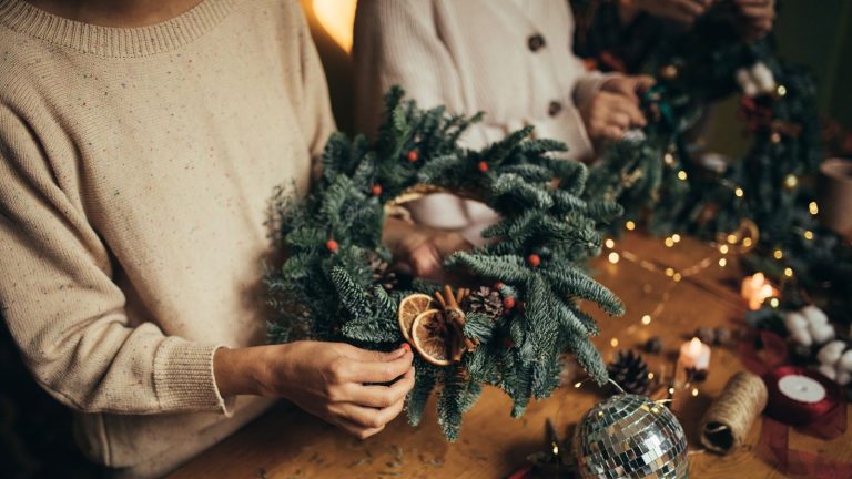 Une couronne de Noël locale est la tendance des fêtes qui est là pour rester – Comment choisir les meilleurs matériaux naturels pour un design de qualité professionnelle Homemade Christmas wreath being held above wooden crafting table