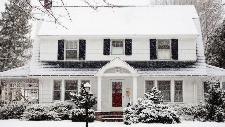 3 règles que les experts en maison suivent toujours pendant les avertissements de neige pour éviter les dommages et les réparations coûteuses A white painted house surrounded by plants, everything covered in snow