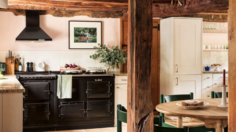 Architecture de caractère, décor en couches et ajouts festifs se combinent pour créer la cuisine la plus confortable de cette maison de campagne. A cozy cottage kitchen with wooden ceiling beams, pink walls, white cabinets, a black AGA, and subtle Christmas decor