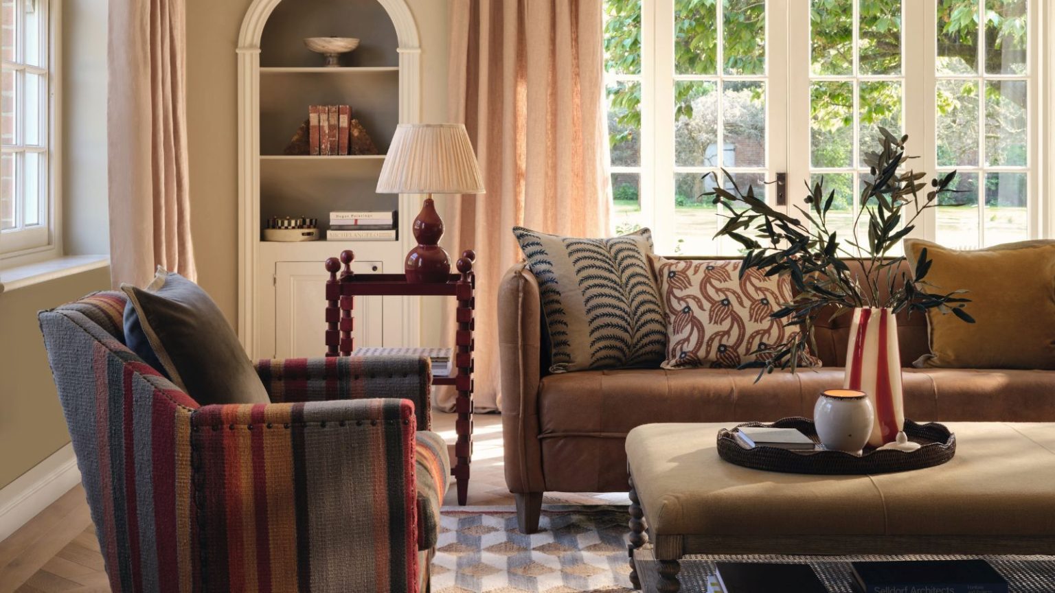 J’ai essayé la méthode simple de désencombrement du panier à linge pour préserver ma santé mentale lors de l’hébergement – L’organisation préalable aux invités est désormais simple et gérable. A warm-toned living room, with a brown three seater sofa and a striped arm chair. An upholstered ottoman table between them, and large glass patio doors behind.